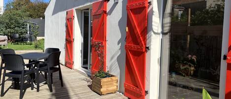 Un patio con una mesa y sillas, una jardinera de madera y contraventanas rojas en un edificio blanco.