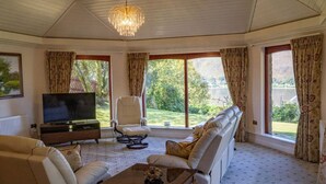 Ein geräumiges Wohnzimmer mit großem Fernseher, bequemen Sitzmöglichkeiten und großen Fenstern mit Blick ins Freie.