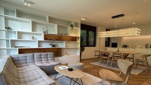 Een moderne woonkamer met een hoekbank, een eethoek met een houten tafel en stoelen, en inbouwkasten met decoratieve objecten.