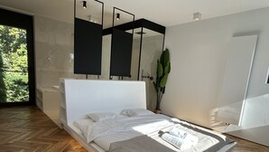Een moderne slaapkamer met een houten visgraatvloer, een wit bed met grijze beddengoed en een grote spiegel boven het bed.