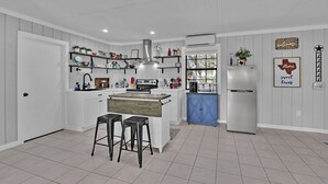 A modern kitchen with white cabinetry, a central island with stools, a stainless steel range hood, and a refrigerator. - Charming Bulverde Farmhouse | Near San Antonio, Patio, BBQ, Hill Views (Bulverde)