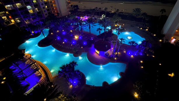 Vista nocturna de un área de piscina de hotel con varias piscinas, palmeras y un edificio grande al fondo.
