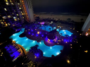 Nachtsicht auf einen Hotelpoolbereich mit mehreren Pools, Palmen und einem großen Gebäude im Hintergrund.