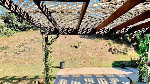 Een overdekt terras met een houten pergola, een betonnen vloer en uitzicht op een grasheuvel.