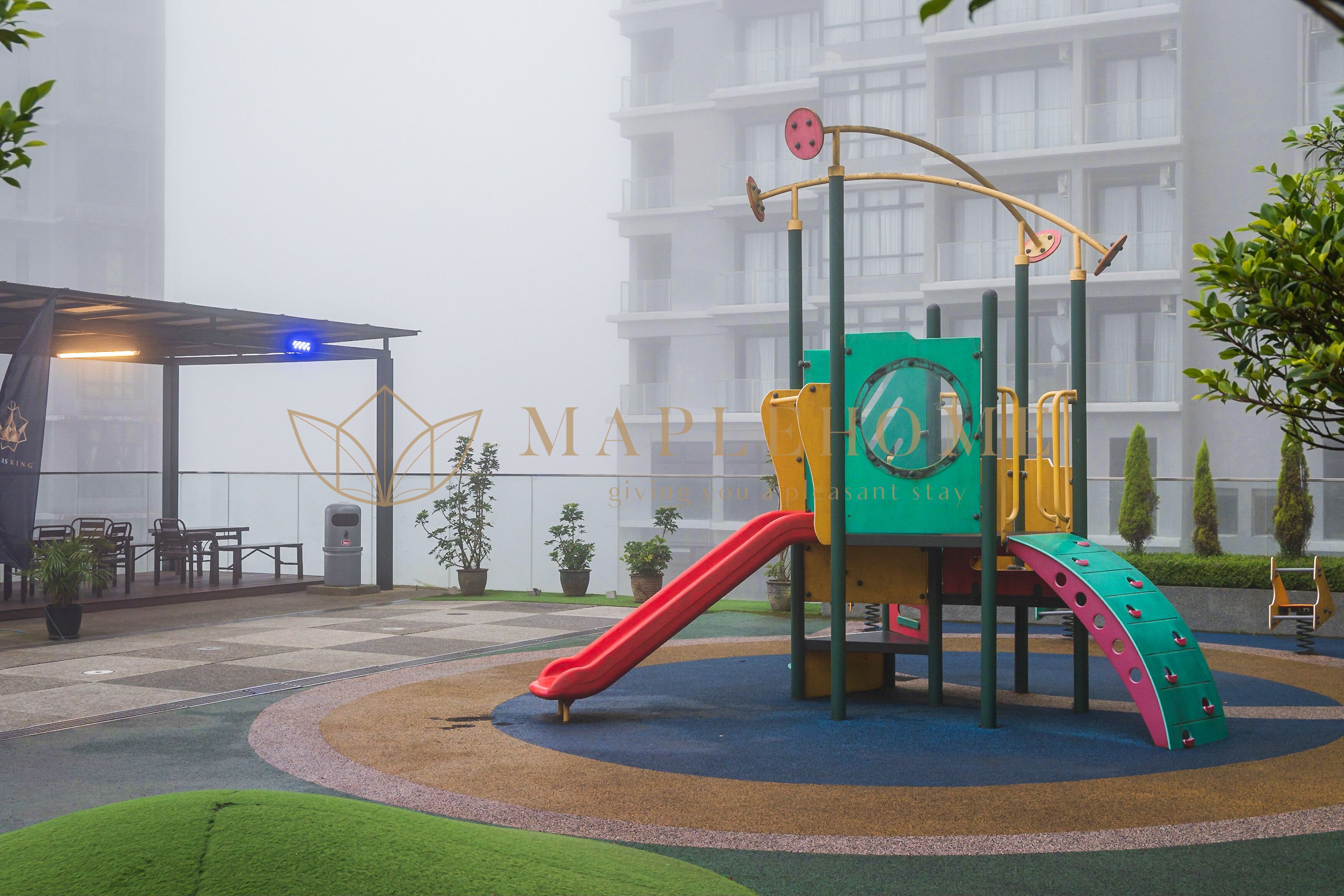 Un parque infantil con tobogán y zona de escalada, rodeado de un jardín ajardinado y un edificio al fondo.
