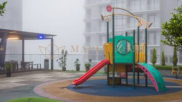 Un parque infantil con tobogán y zona de escalada, rodeado de un jardín ajardinado y un edificio al fondo.