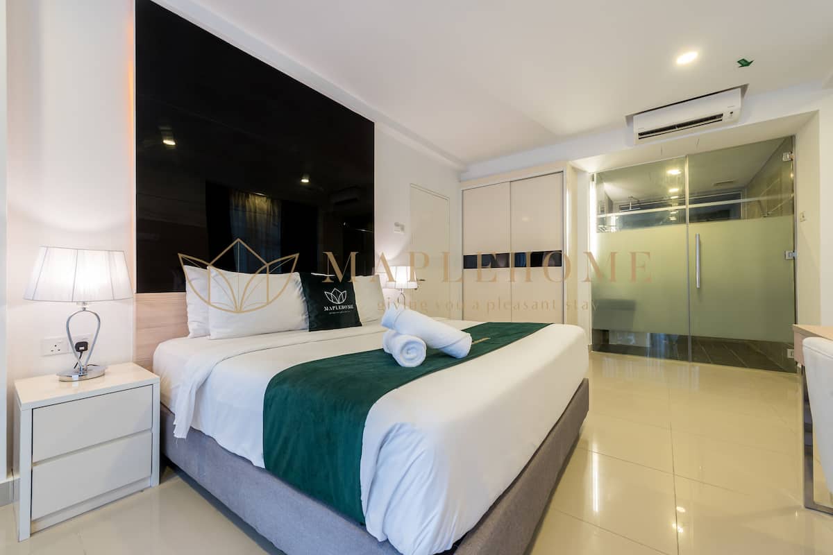 A hotel room with a neatly made bed, a nightstand with a lamp, a green throw blanket, and a glass shower area.