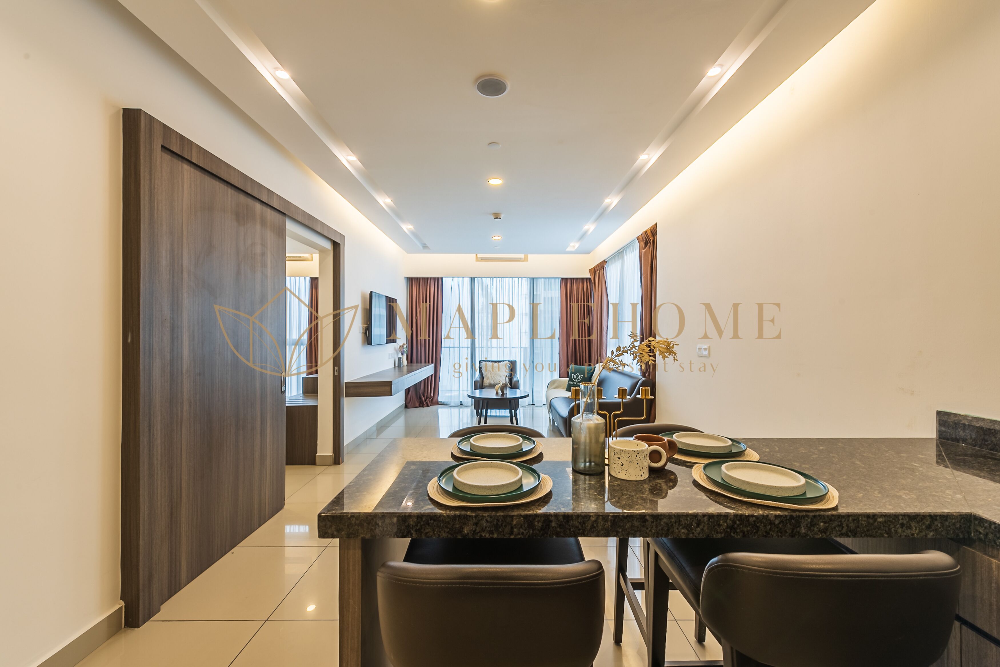 A modern dining area with a long table set for four, a kitchen in the background, and a view of the living area.