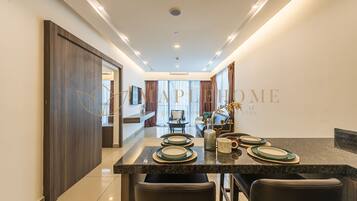 A modern dining area with a long table set for four, a kitchen in the background, and a view of the living area.