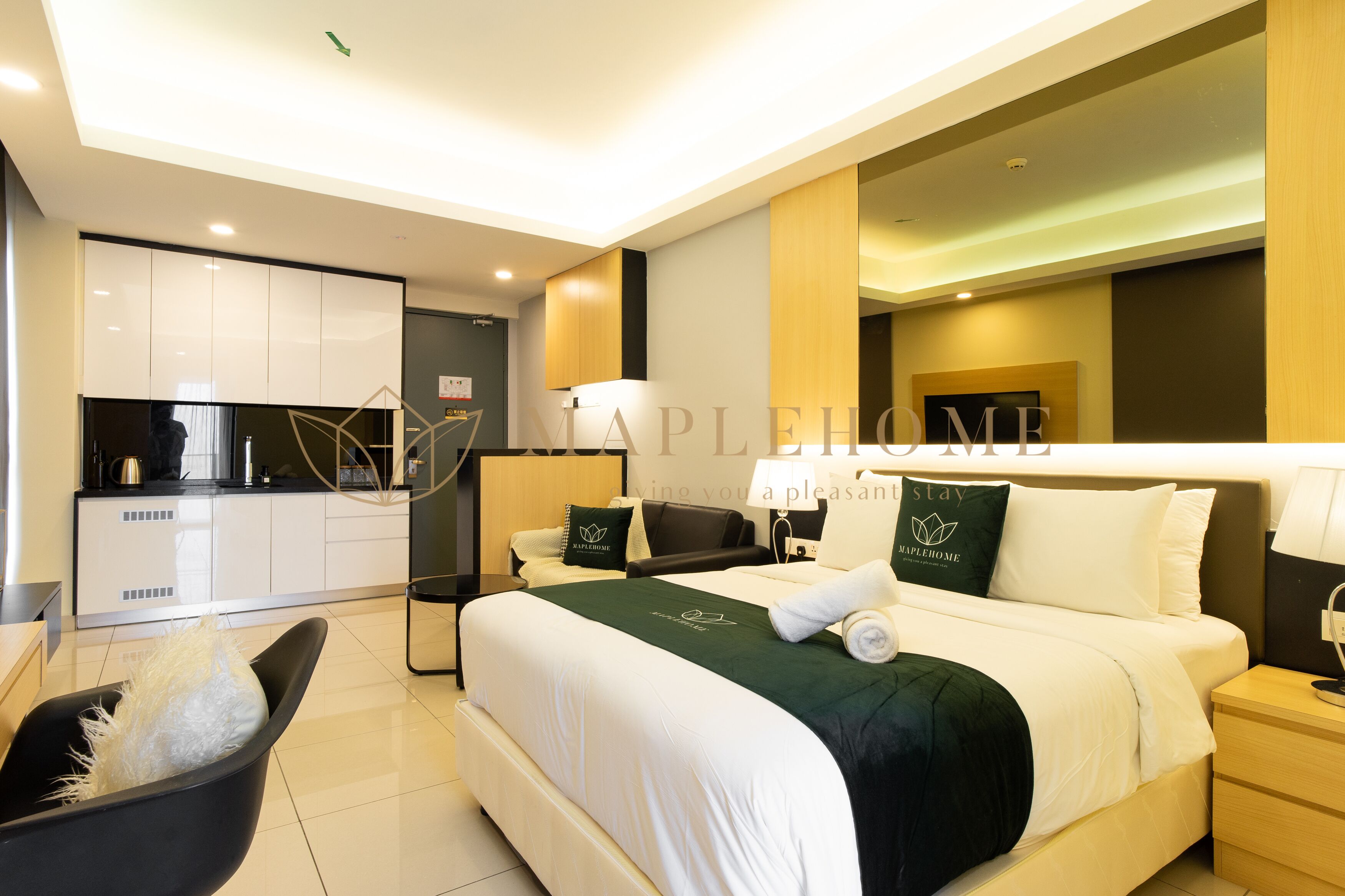 A modern hotel room with a neatly made bed, a small bedside table, a chair, and a kitchenette in the background.