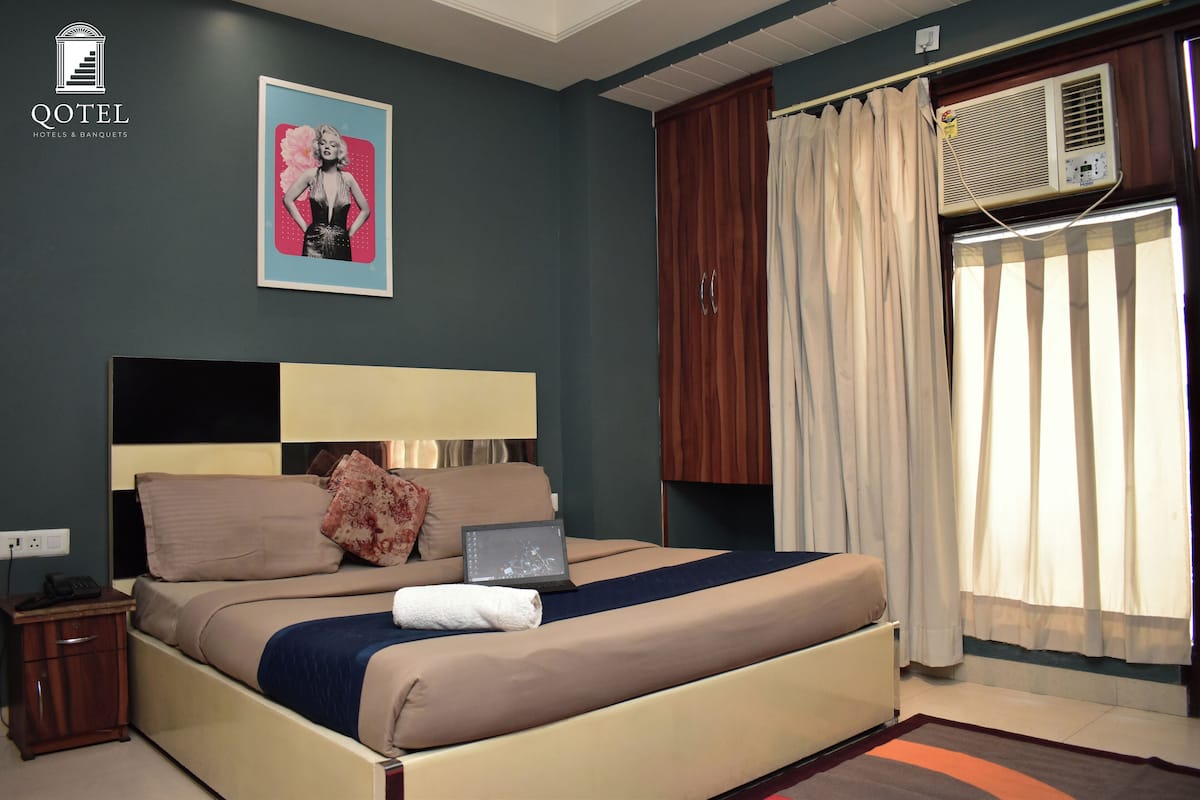 A hotel room with a bed, a bedside table with a lamp, a poster on the wall, and an air conditioning unit.