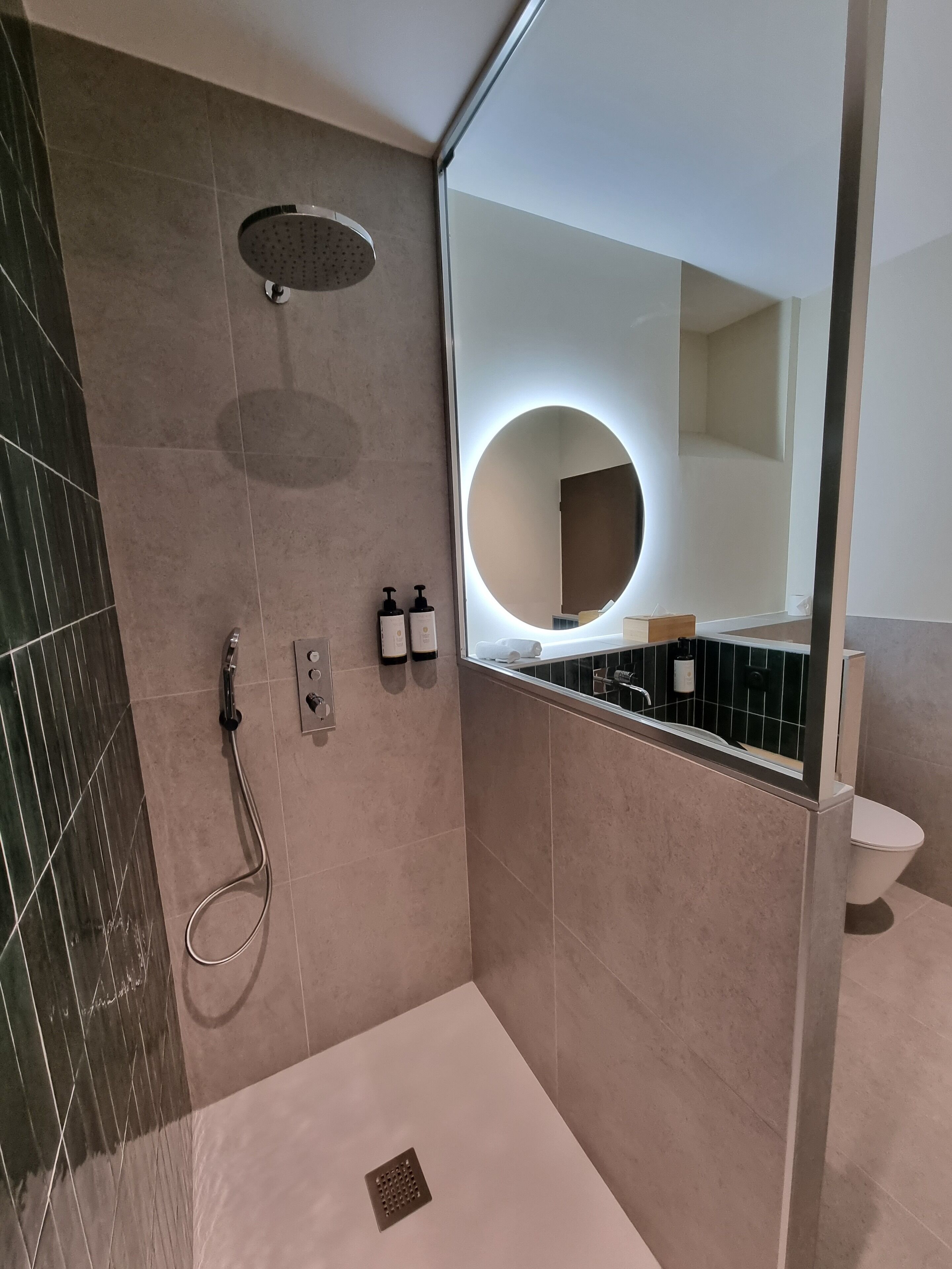 Une salle de bain moderne équipée d’une douche, de toilettes et d’un grand miroir avec un éclairage circulaire.