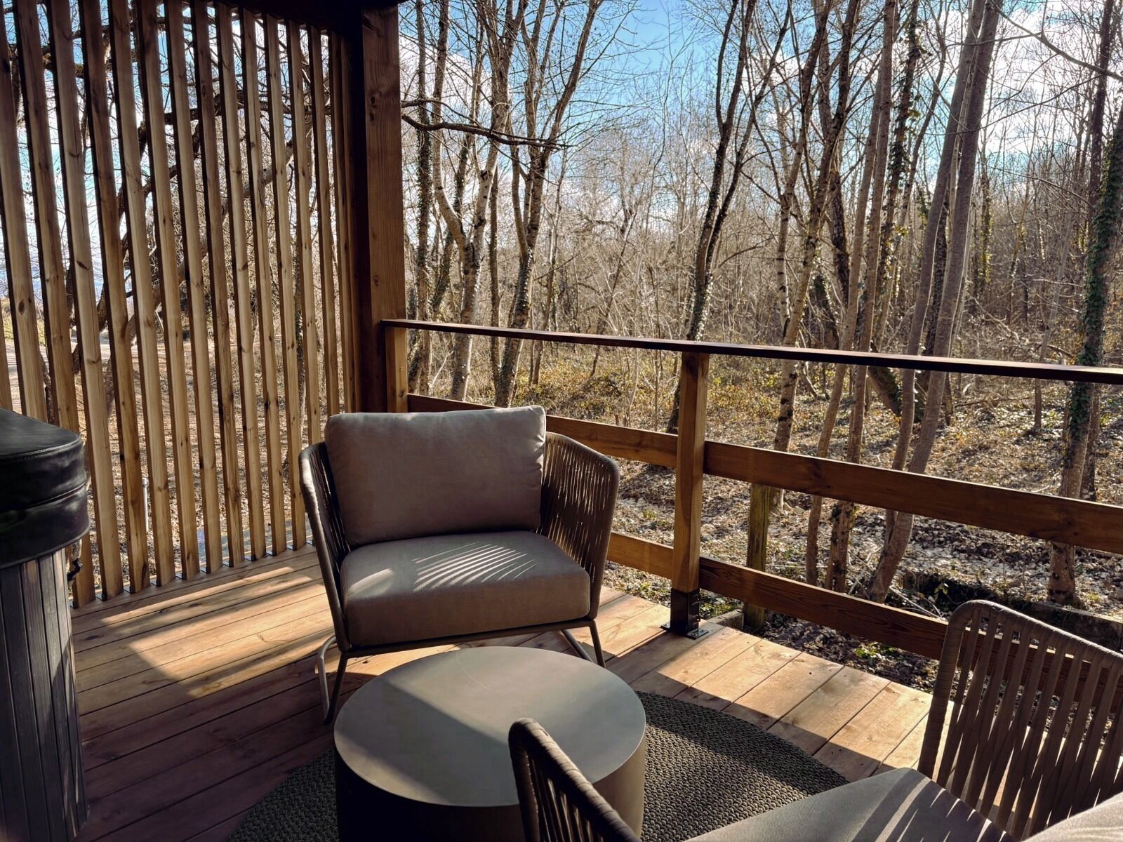 A wooden deck with a chair and table, surrounded by a wooden railing, overlooking a forested area.