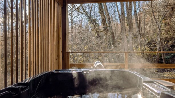 Un jacuzzi con vapor que se eleva, rodeado de barandas de madera y vistas a un bosque.