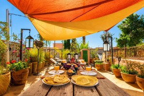 An outdoor dining area with a wooden table, potted plants, and a large orange canopy.
