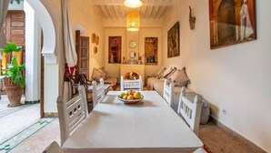 A dining area with a long table, white chairs, a chandelier, and a bowl of fruit. - Riad Dar Al Barakaa - Exclusive- Breakfast (Marrakech)