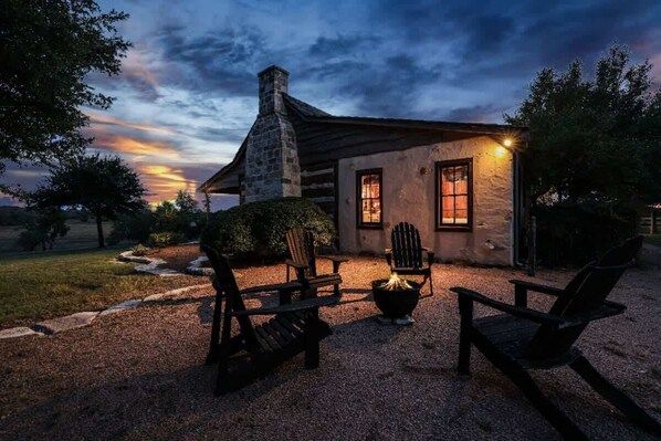 A rustic cabin with a stone chimney, wooden exterior, and a gravel patio with Adirondack chairs.
