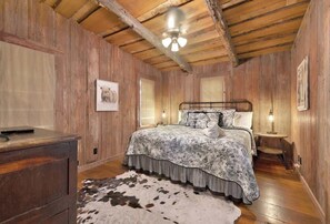 A rustic bedroom with wooden walls, a bed with a floral bedspread, a ceiling fan, and a cowhide rug. - Private Ranch Suite-Bar-GameRoom (Fredericksburg)