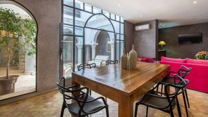 A dining area with a wooden table and chairs, a pink sofa, and a TV mounted on the wall. - Riad Princesses des Sables-Jaccuzzi-Breakfast (Marrakesh)