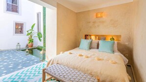 A bedroom with a bed, bedside table, and a bench. There is a window and a door leading to an outdoor area. - Riad Itri (إتري) Exclusif - Marrocan Breakfast (Marrakesh)