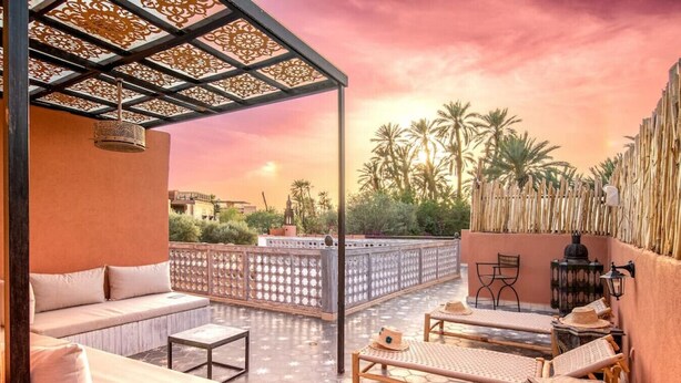 A rooftop terrace with a sofa, a wooden table, and a chair, overlooking palm trees and a sunset. - Riad Itri (إتري) Exclusif - Marrocan Breakfast (Marrakesh)