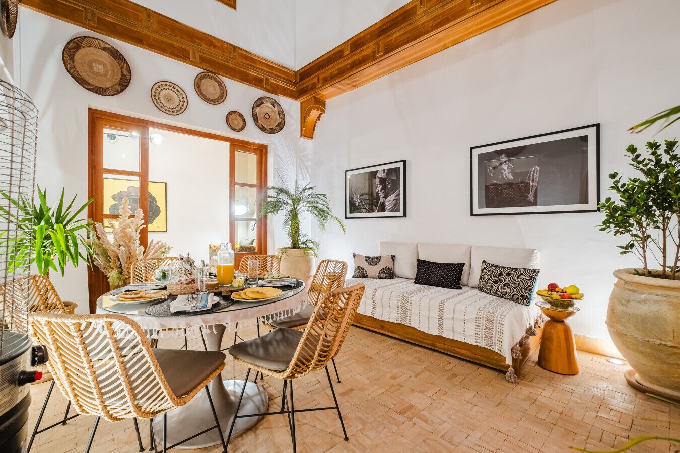 Un comedor con sillas de mimbre, una mesa redonda y una barra de buffet. Hay un sofá con cojines estampados y una planta grande en maceta.