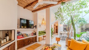 A spacious living area with a flat-screen TV, bookshelves, a green sofa, a coffee table with magazines and fruit, and a dining area with a table and chairs. - Riad Ouladi -Exclusif - 5mn Jemaa El Fnaa -Pool (Marrakesh)