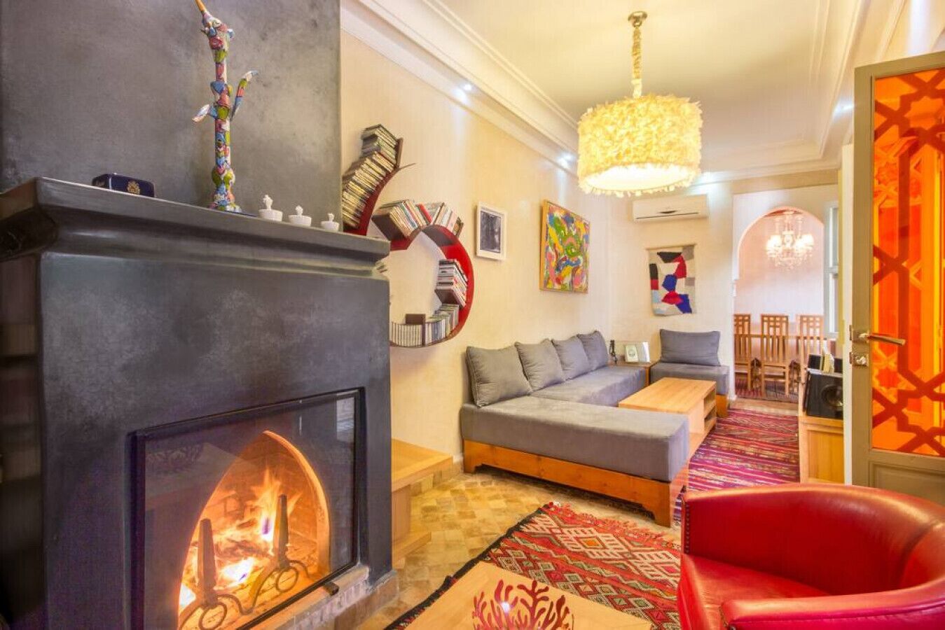 Un salon chaleureux avec une cheminée, un canapé, un fauteuil rouge et, au fond, un coin repas.