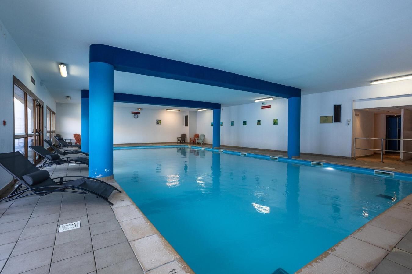 Een binnenzwembad met blauwe pilaren, lounge fauteuils en een betegelde vloer.