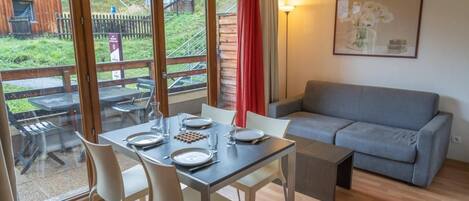 A room with a dining table set for four, a grey sofa, and a large window with a view of a wooden building and a fence.