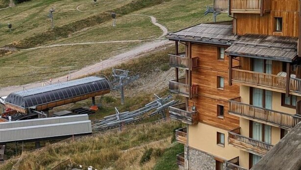 Un edificio in legno a più piani con balconi, sullo sfondo di un paesaggio collinare e una funivia da sci.