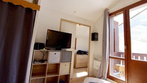 Quarto com televisão sobre um armário de madeira, janela com vista para o exterior e porta que dá para outro cômodo.