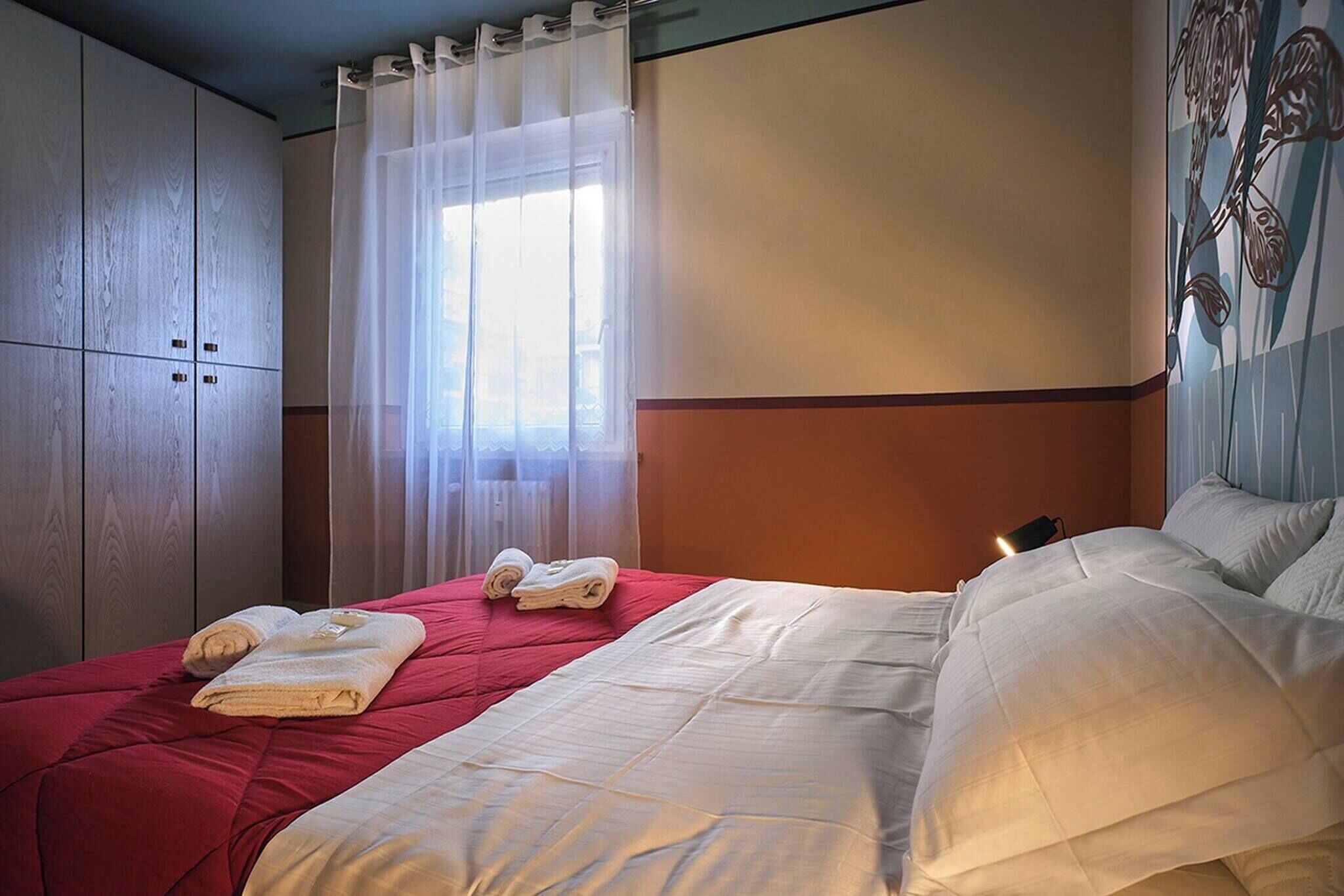 Une chambre d’hôtel avec un lit, une fenêtre avec des rideaux légers, un radiateur et un mur orné d’un motif floral.
