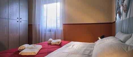 A hotel room with a bed, a window with sheer curtains, a radiator, and a wall with a floral pattern.