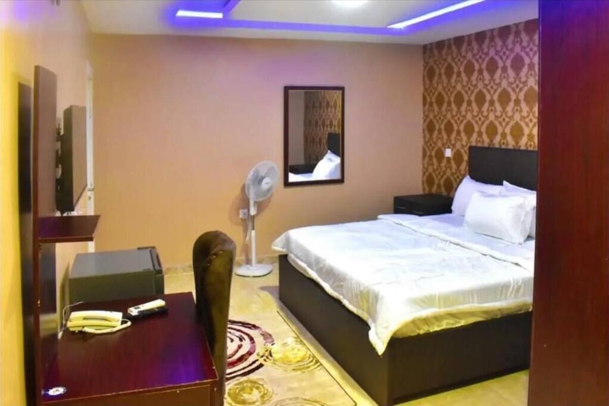 Habitació d'hotel amb llit, taula de nit, cadira, escriptori i ventilador de peu.