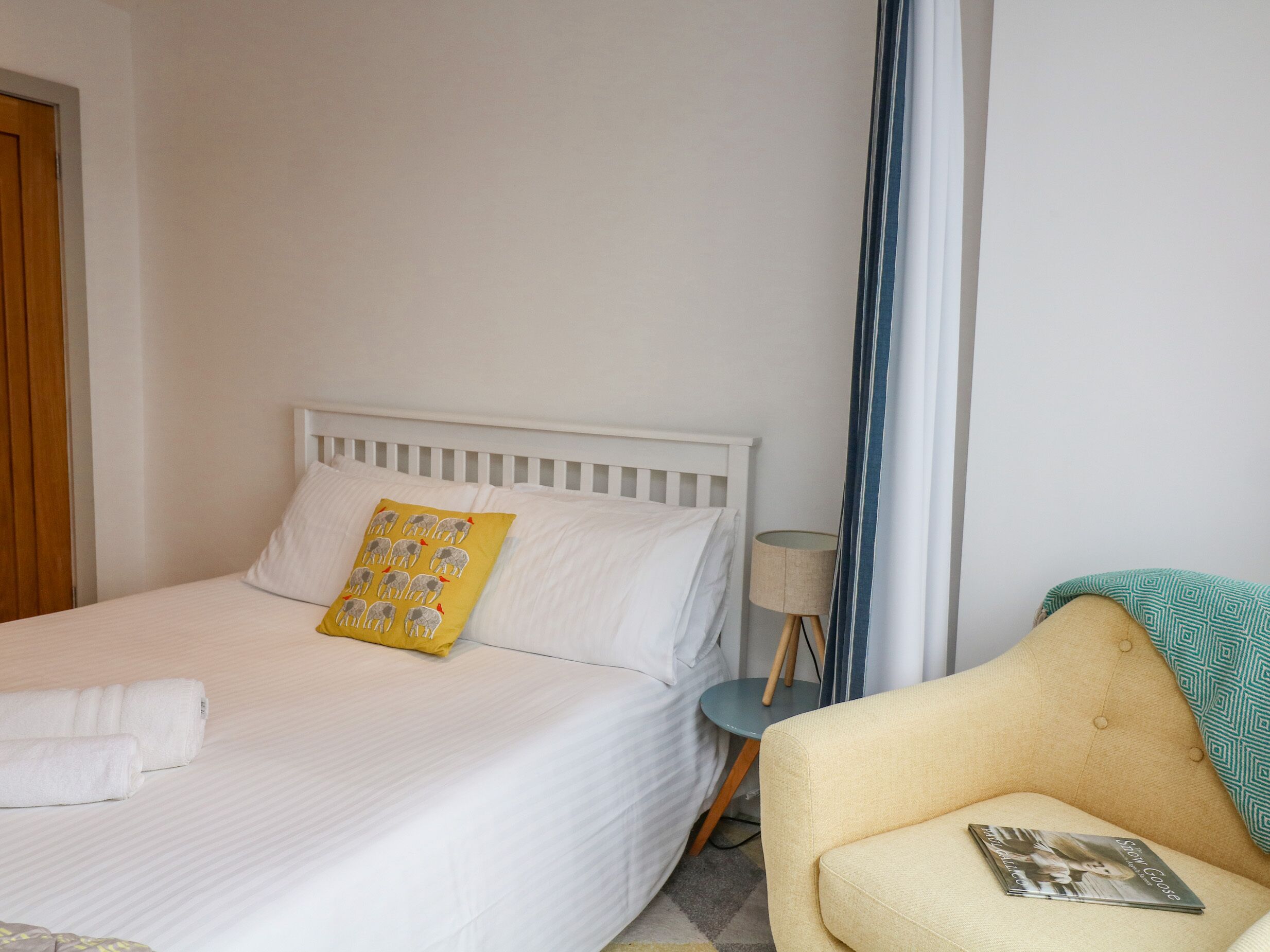 A neatly made bed with white linens and a yellow pillow, a small bedside table with a lamp, and a sofa with a magazine on it.
