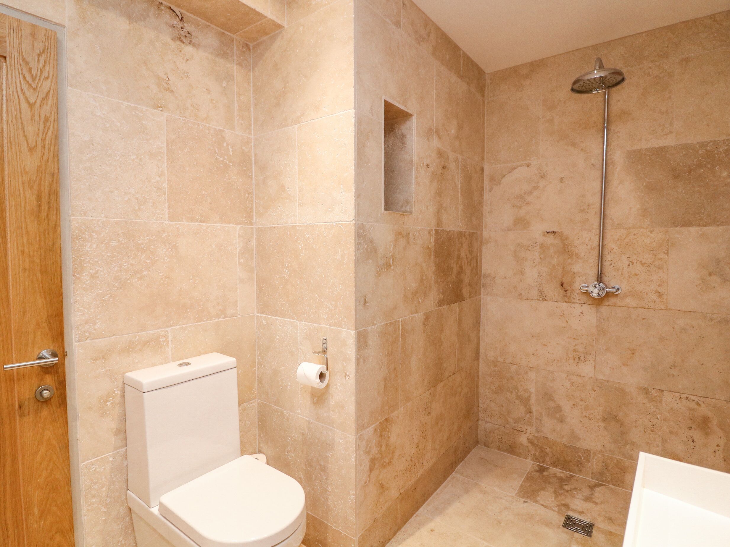 A bathroom with beige tiles, a toilet, a shower with a handheld showerhead, and a small window.