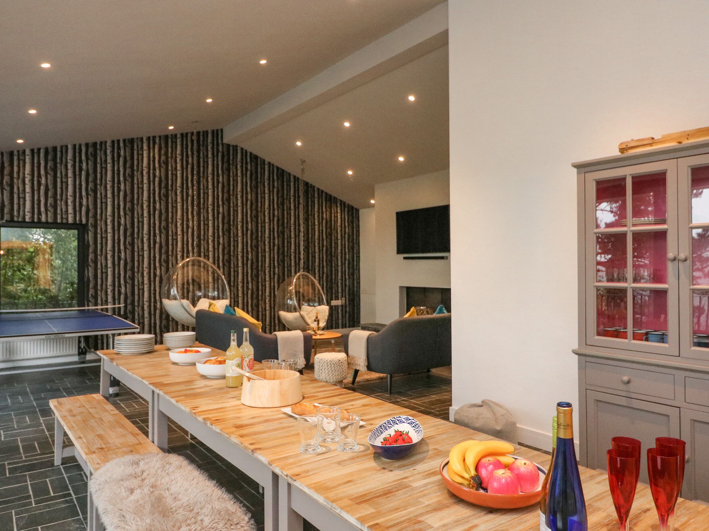 A modern living room with a ping pong table, seating area with chairs, a dining table with food and drinks, and a cabinet with glass doors.