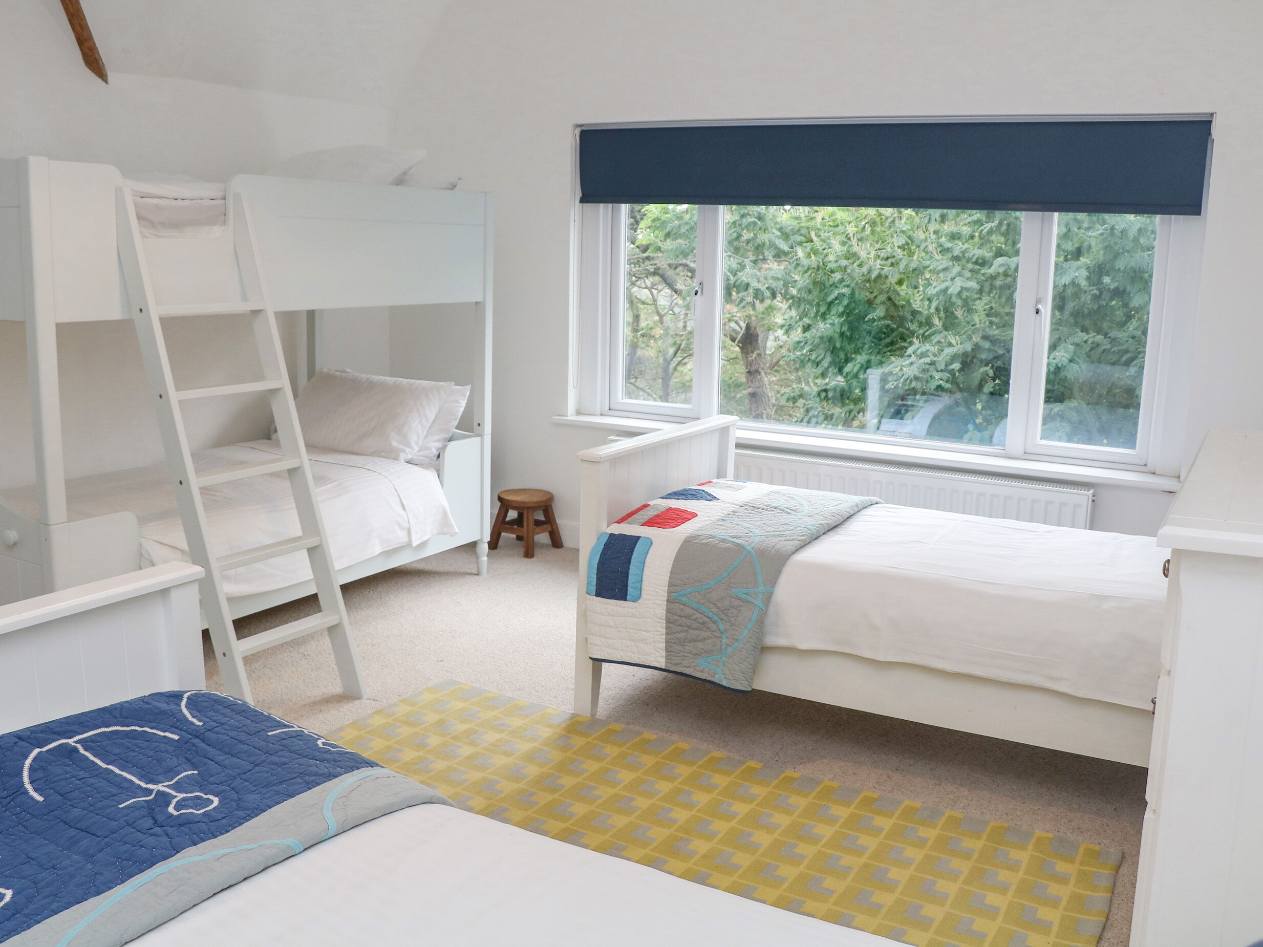 Une chambre d’enfant avec des lits superposés, un petit tabouret et une fenêtre donnant sur un paysage verdoyant.