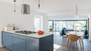 A modern kitchen with a central island, a dining area with a large table and chairs, and a view of the outdoors through large windows. - Landmark (Salcombe)
