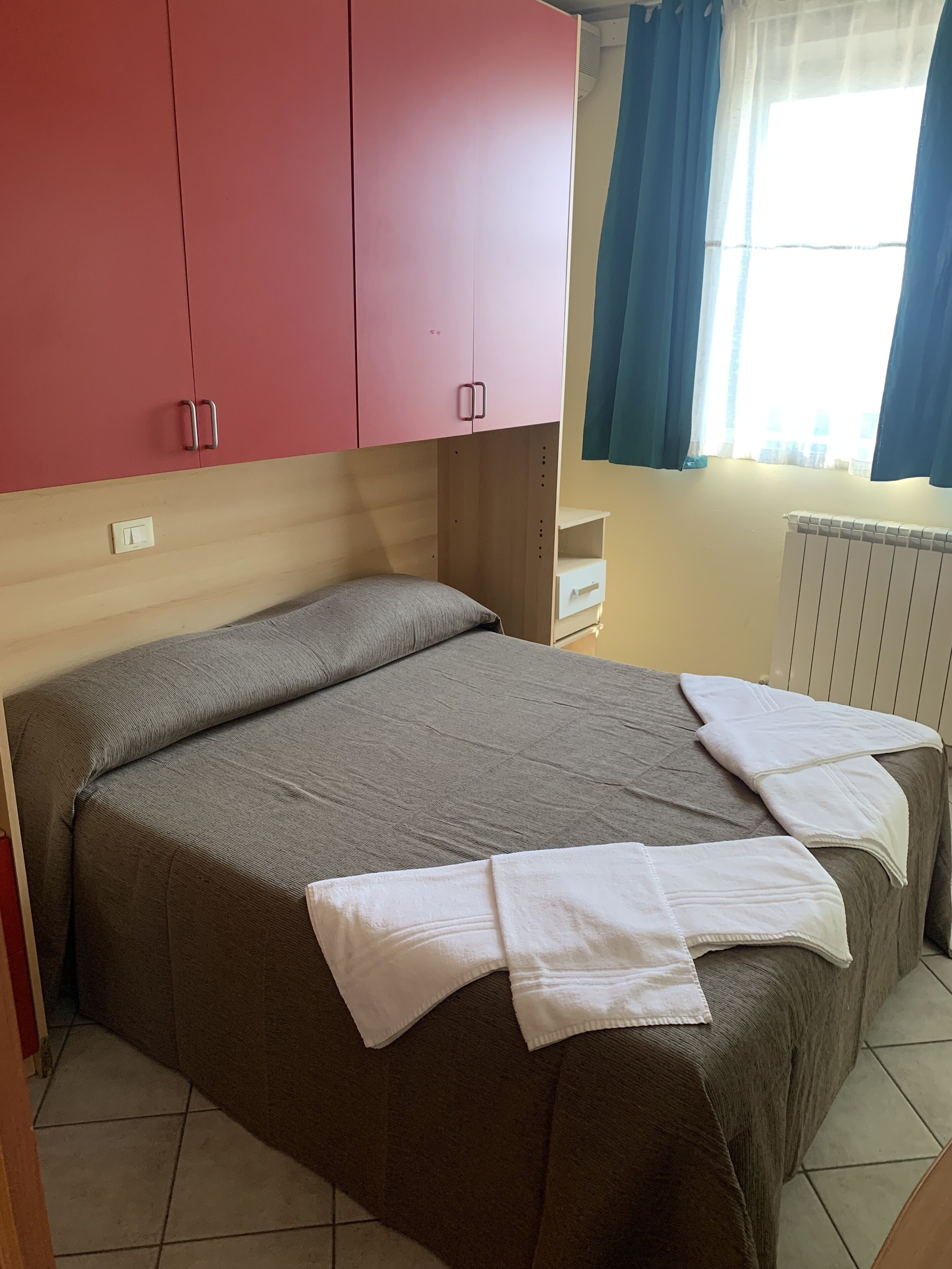 A room with a bed, a red wardrobe, a white bedside table, and a window with blue curtains.