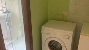 Une petite buanderie équipée d’un lave-linge à chargement frontal, d’un petit meuble blanc et d’une cabine de douche en verre.
