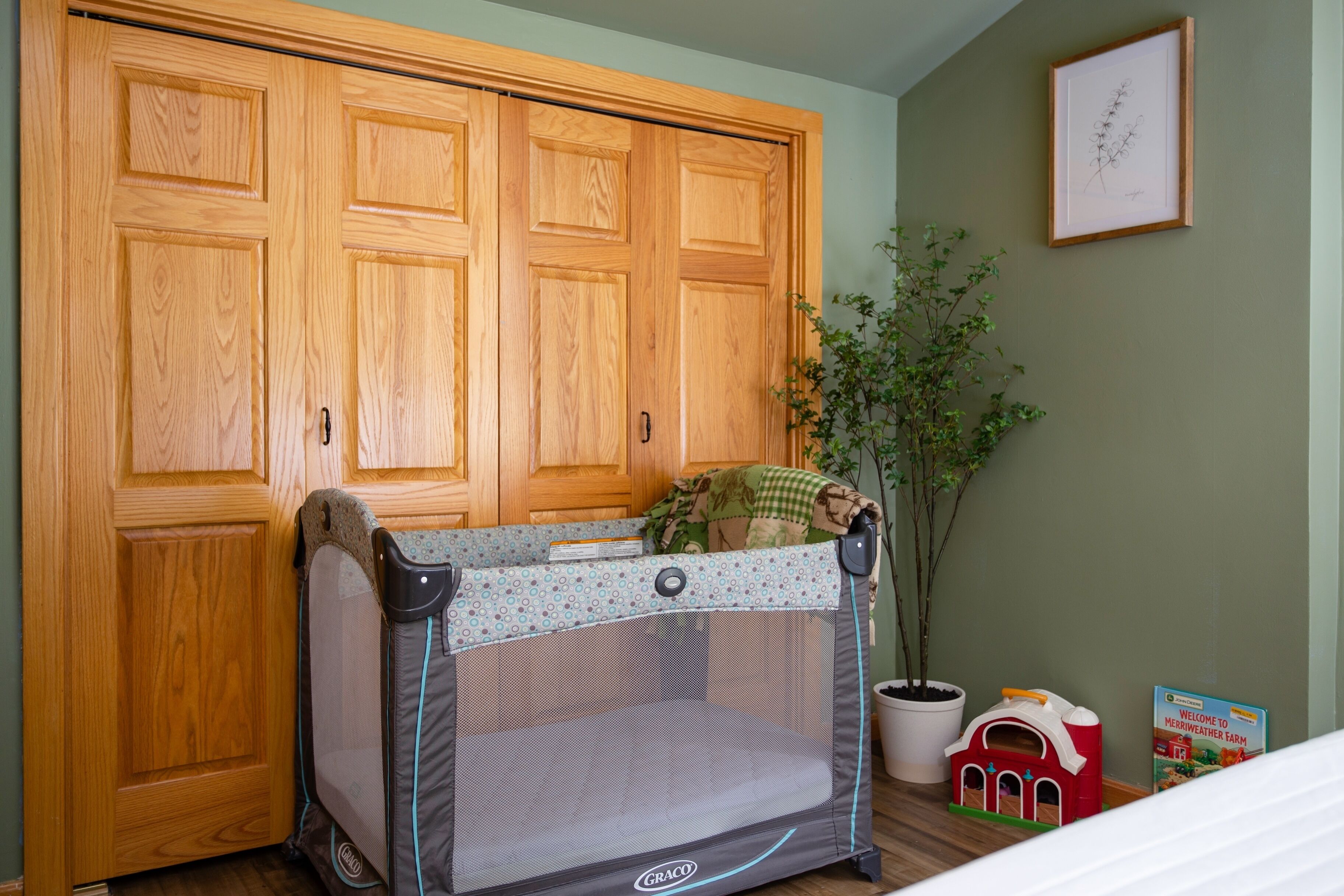 Une pièce comprenant une armoire en bois, un parc pour bébé de la marque Graco, une plante en pot et une maison de jeu.