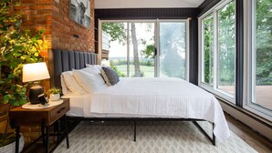 A bedroom with a large bed, a nightstand, a lamp, a potted plant, and a view of greenery through large windows.