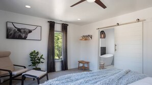 Un dormitorio con cama, una silla, una planta, ventilador de techo, ventana con cortinas y baño con bañera y perchero para toallas.