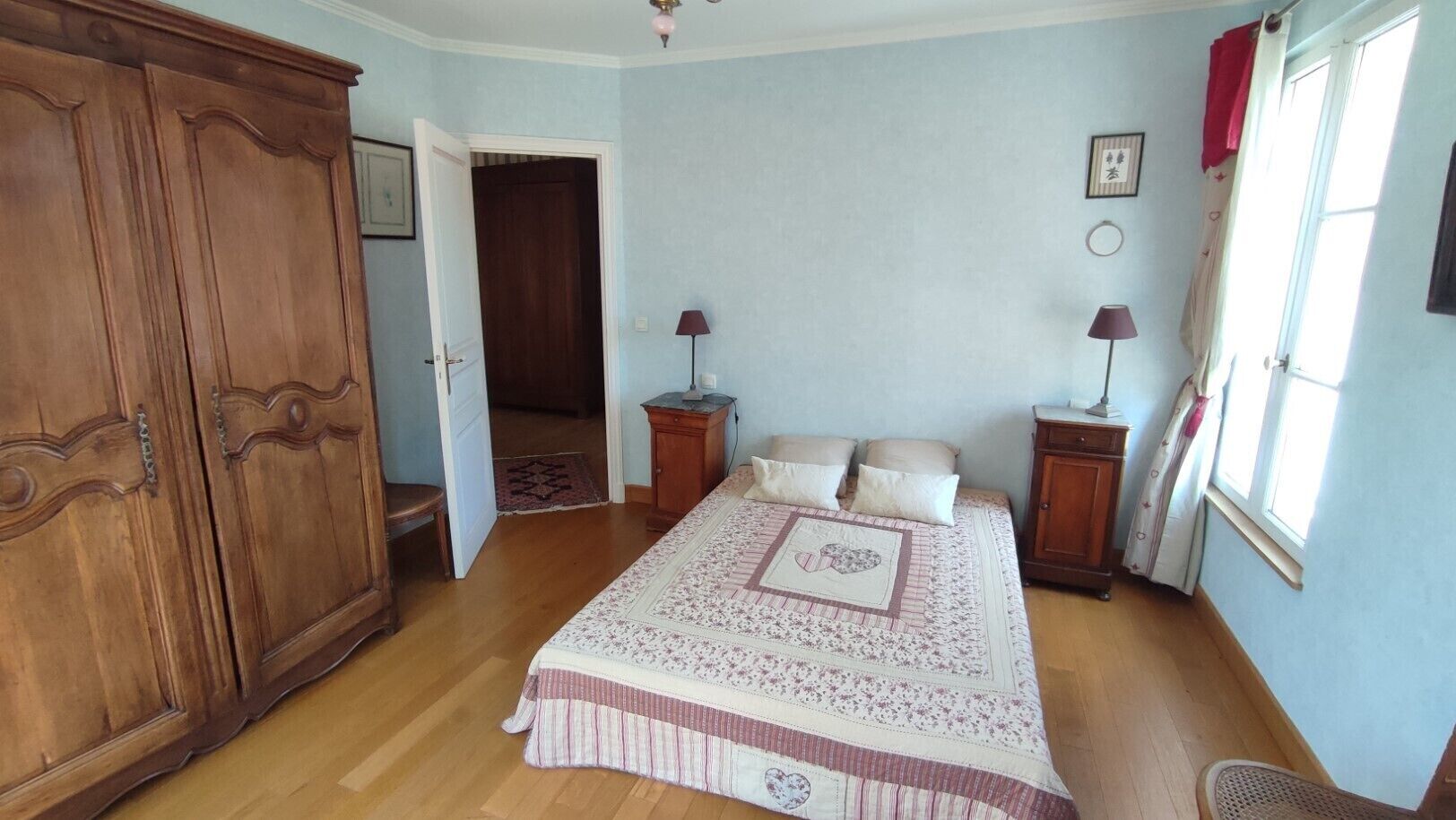 Une chambre avec une grande armoire en bois, un lit recouvert d’une couvre-lit à motifs floraux, deux tables de chevet avec des lampes et une fenêtre avec des rideaux.