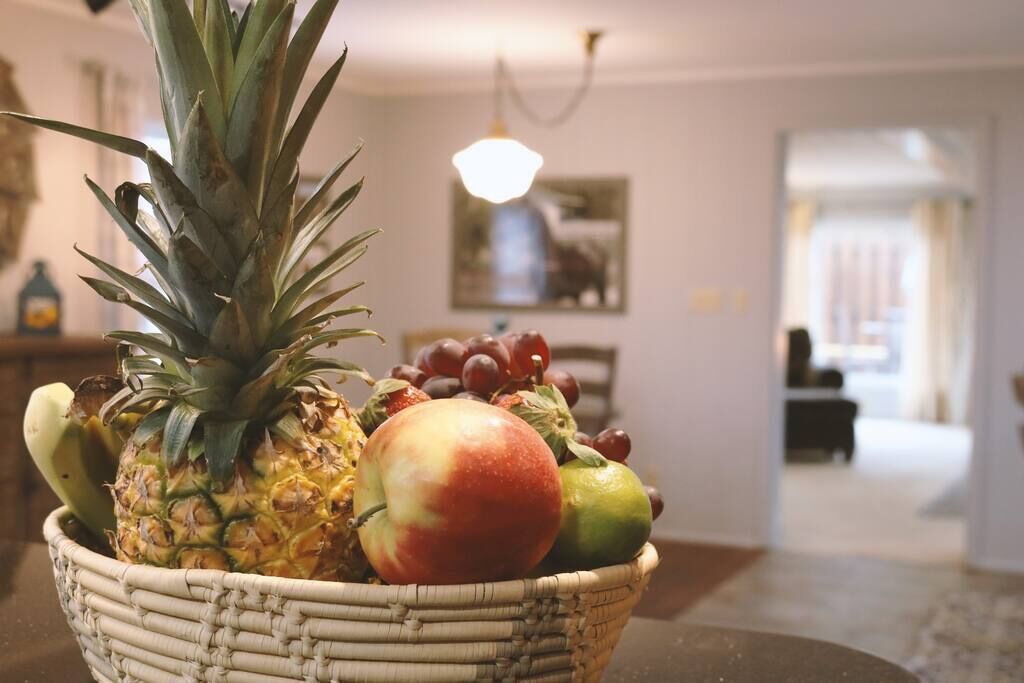Una cesta de mimbre llena de frutas, entre ellas una piña, una manzana y uvas, sobre una mesa en una habitación con una araña y un cuadro en la pared.