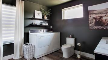 A laundry area with a washer and dryer, a toilet, a trash can, and a window with blinds.