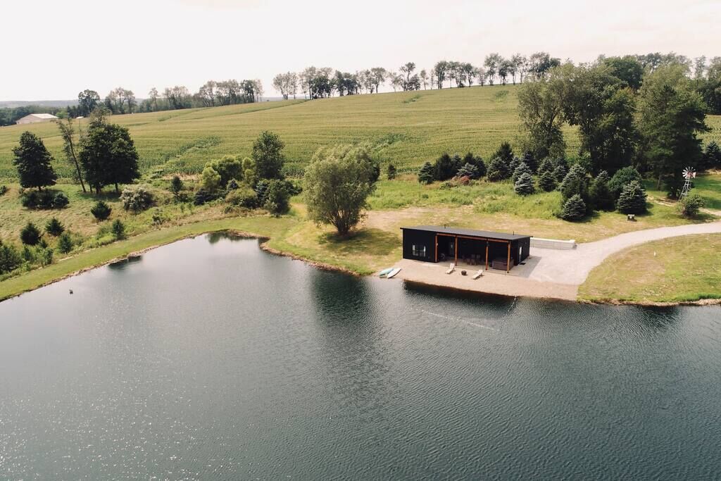 Eine Holzhütte am See mit einer Anlegestelle, umgeben von Bäumen und Feldern.