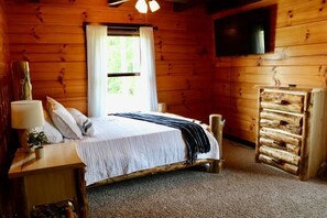 Un dormitorio de cabaña de madera con una cama, una mesita de noche, una lámpara, un televisor montado en la pared y un banco de troncos.